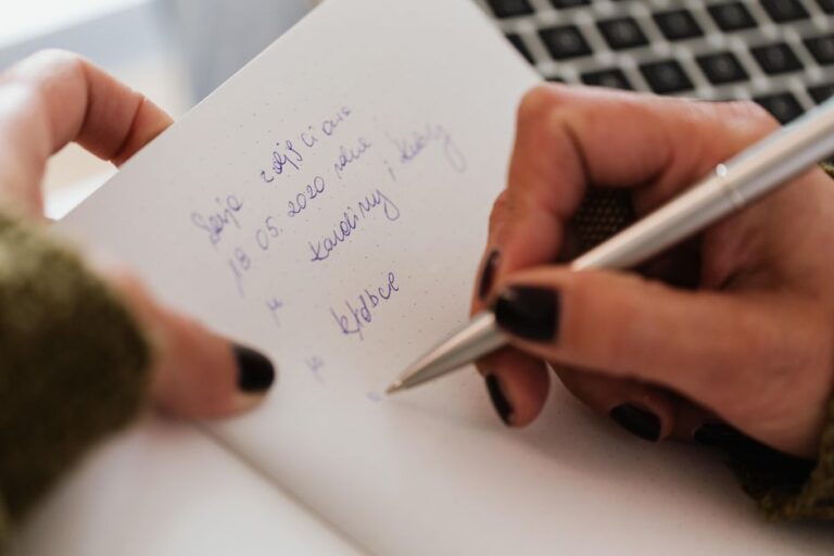 Crop anonymous female with manicured hands writing down notes in notebook with white sheets by using stylish silver pen near modern laptop